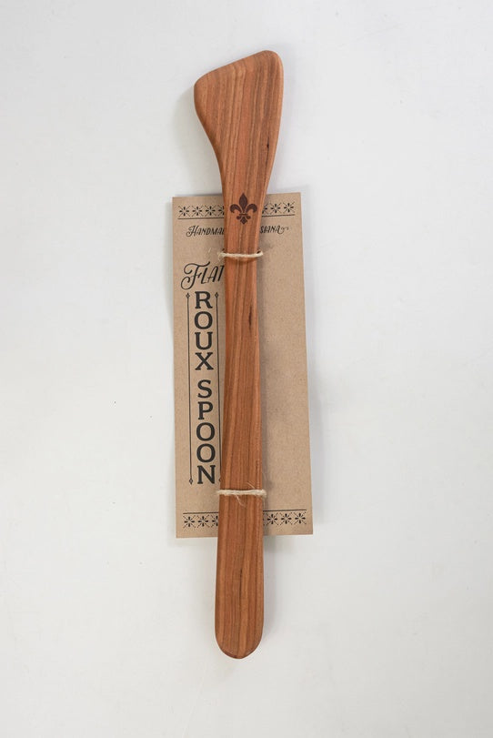 Hand-carved wood flat roux spoon handmade in Louisiana features an asymmetrical paddle head engraved with a fleur-de-lis emblem and natural reddish-brown grain secured to a kraft paper tag with rustic twine against a neutral white background