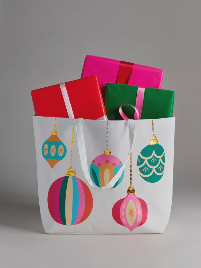 Studio photograph of a white tote bag filled with wrapped gifts in a studio setting, featuring colorful retro ornament illustrations in teal and pink alongside vibrant green and fuchsia gift boxes with contrasting ribbons