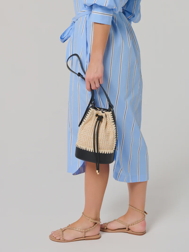 Studio fashion photograph of a woman in a neutral studio holding a cream crocheted bucket bag with black leather trim, wearing a light blue striped dress and gold-studded tan sandals