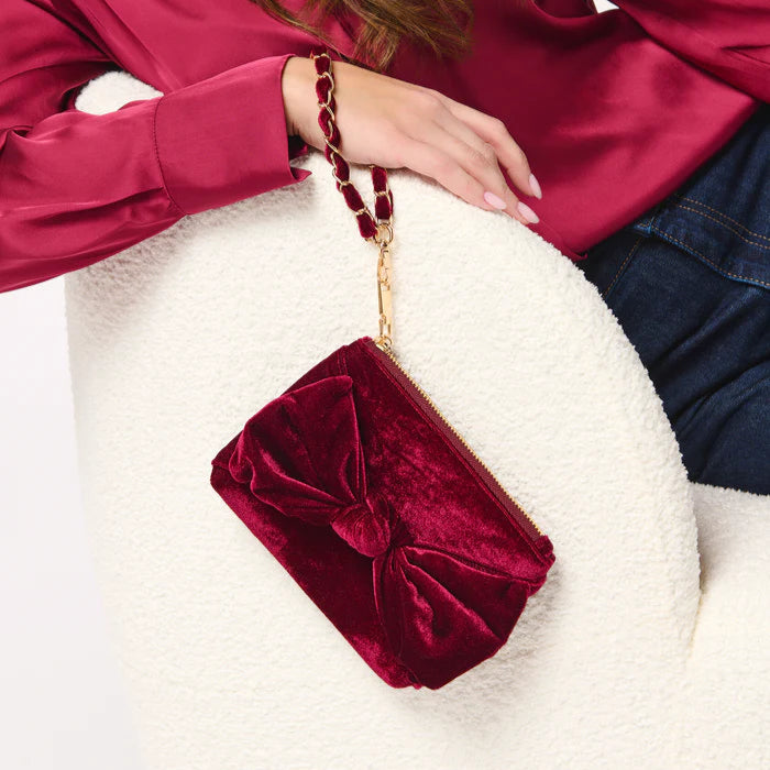 Fashion photograph of a burgundy velvet bow clutch bag with a gold chain strap held by a person wearing a magenta satin blouse while sitting in a white textured bouclé armchair