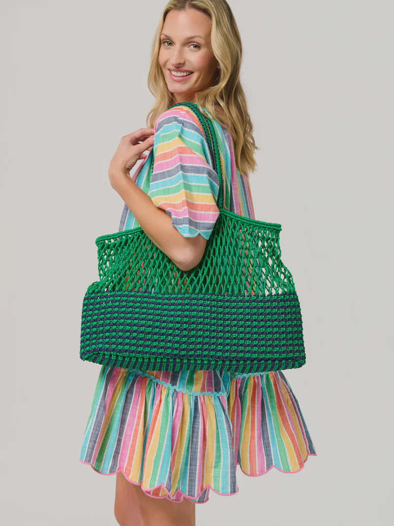 Studio photograph of a smiling blonde woman in a studio carrying a large green and navy blue macramé tote bag while wearing a vibrant multi-colored striped dress with a scalloped hem