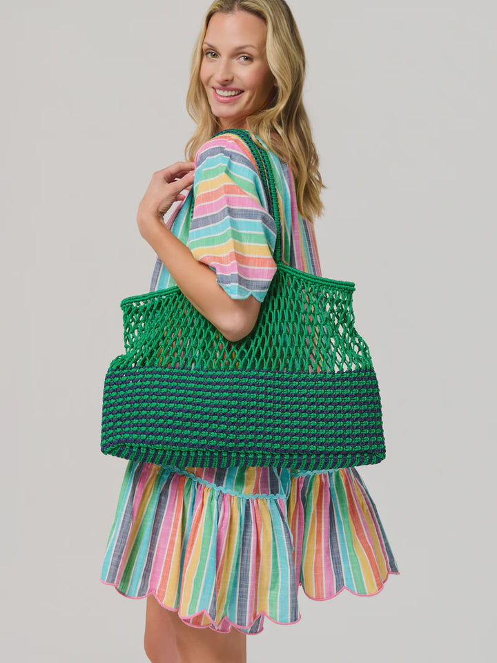 Studio photograph of a smiling blonde woman in a studio carrying a large green and navy blue macramé tote bag while wearing a vibrant multi-colored striped dress with a scalloped hem