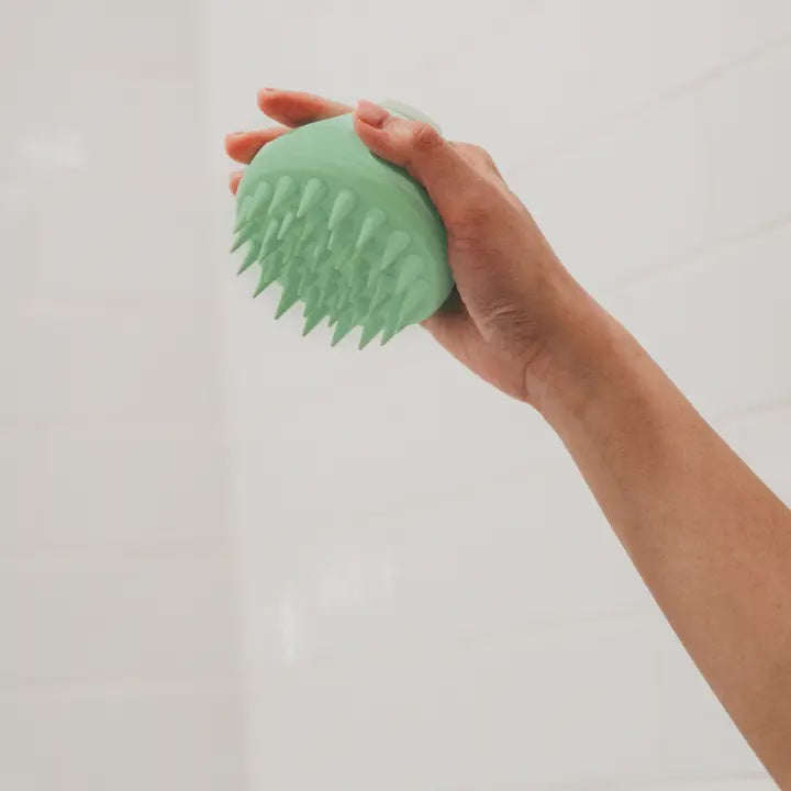 Photograph of a person's hand holding a mint green silicone scalp massager with tapered bristles against a background of white bathroom tiles