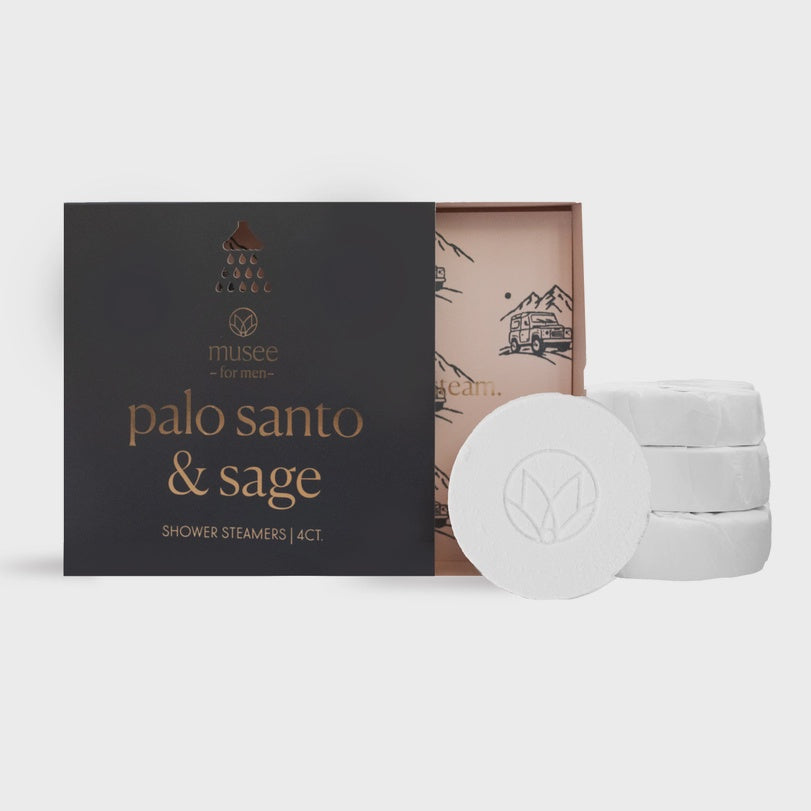 Product photograph by Musee of palo santo and sage shower steamers for men featuring a charcoal gray box with copper foil typography, white tablets with an embossed floral logo, and a peach interior illustrated with a mountain landscape and vintage off-road vehicle