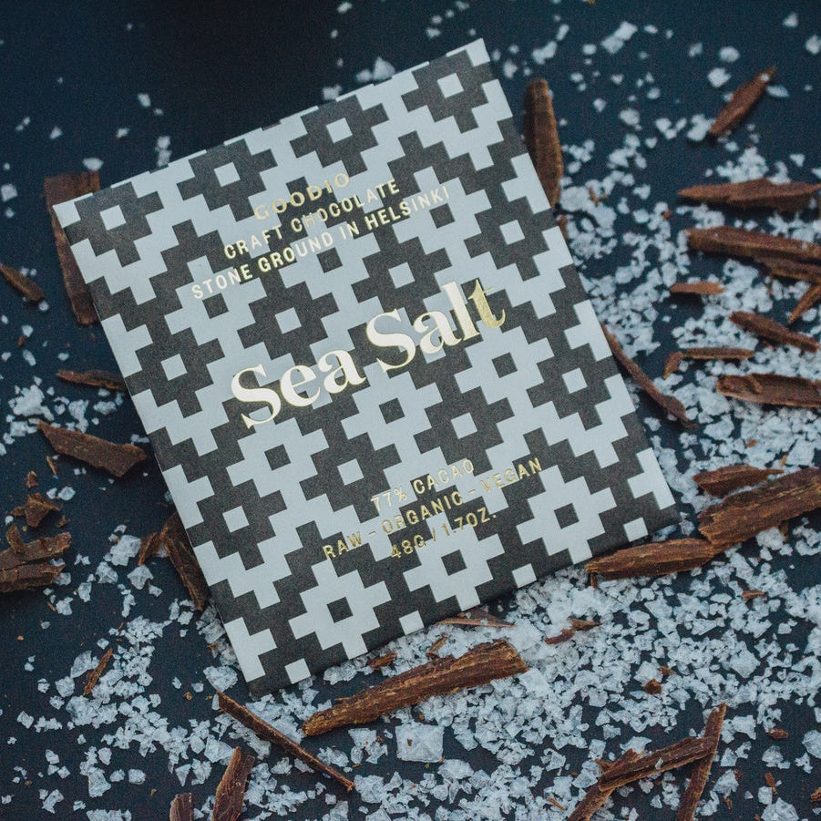 Product photograph of Goodio Craft Chocolate from Helsinki featuring a Sea Salt bar with a black and white geometric cross pattern surrounded by scattered coarse sea salt crystals and dark chocolate shavings