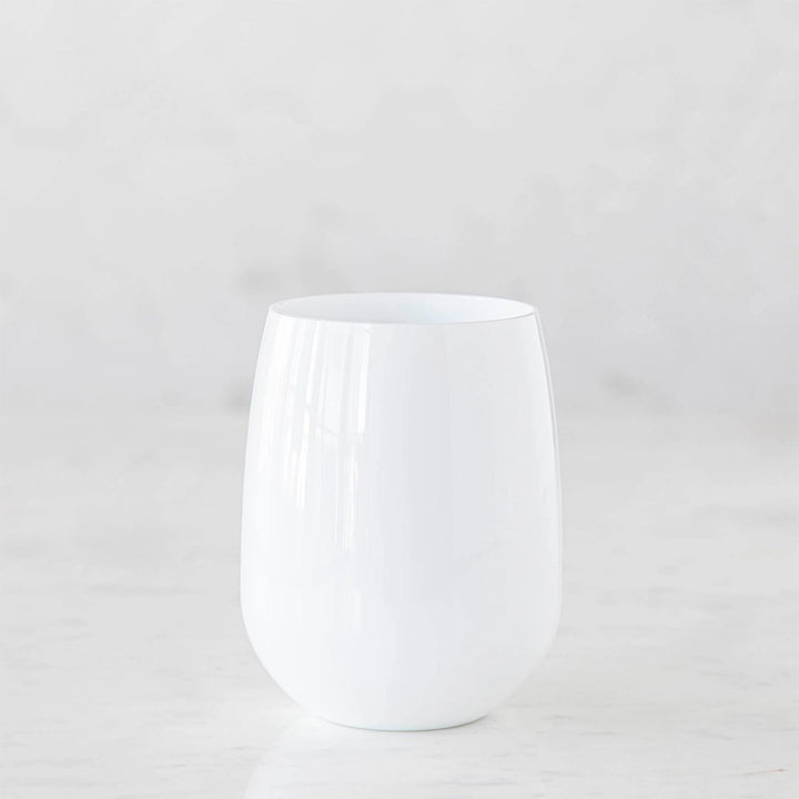 Studio photograph of a stemless opaque white glass tumbler on a white marble surface featuring a smooth curved silhouette and subtle vertical light reflections against a minimalist background