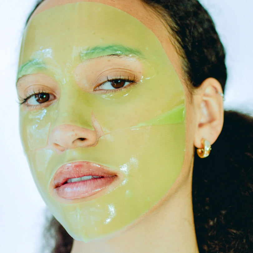 Photograph of a woman in a studio setting wearing a translucent lime-green gel face mask with a small gold hoop earring and dark curly hair