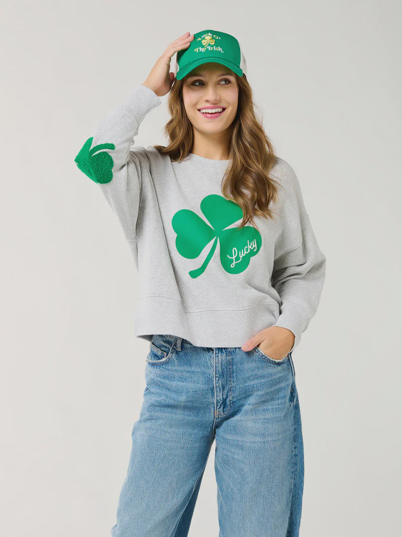 Studio photograph of a smiling woman in a studio wearing a grey sweatshirt featuring a large green shamrock graphic, textured clover elbow patches, and a green Luck of the Irish trucker hat