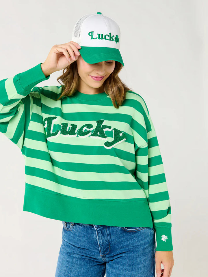Studio photograph of a woman wearing a green and light green striped sweatshirt with textured Lucky lettering, a matching white and green trucker hat featuring a small shamrock, and blue denim jeans against a plain white background