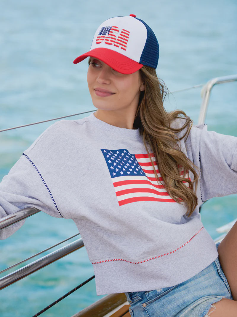 Photograph of a woman on a sailboat wearing a patriotic USA trucker hat and a grey sweatshirt featuring a large American flag graphic and decorative red and white braided rope accents