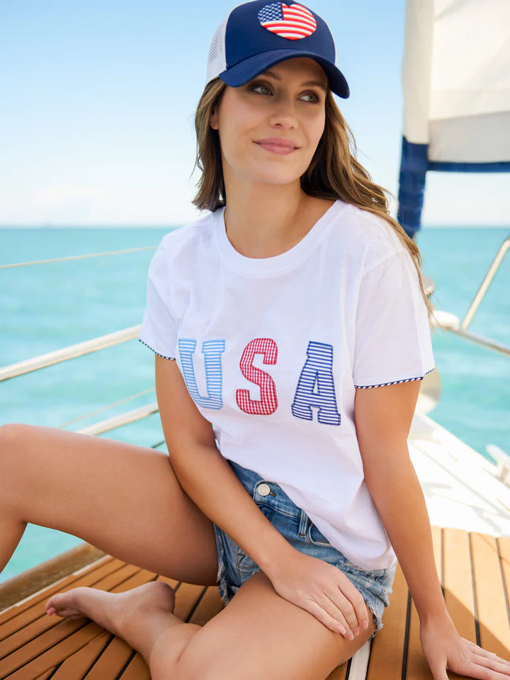 Photograph of a woman sitting on the teak deck of a sailboat on the open water wearing a white t-shirt with red and blue USA patterned letters and a navy hat with a heart-shaped American flag patch