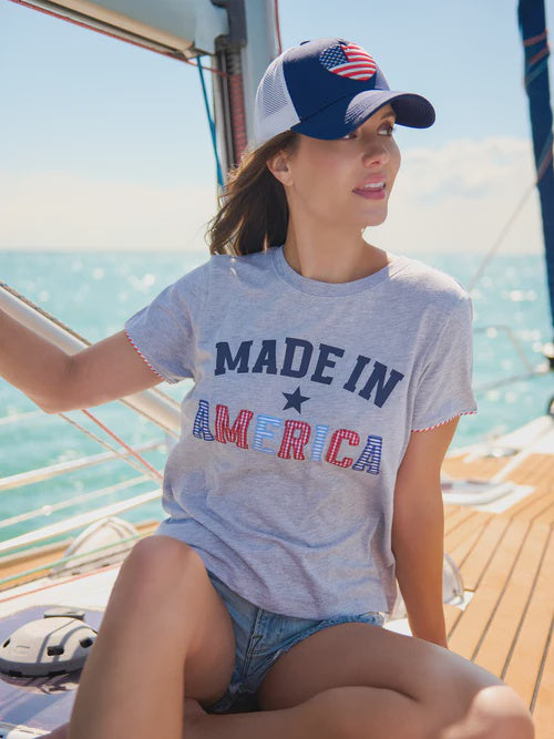 Photograph of a woman on a wooden sailboat deck at sea wearing a heathered grey Made in America t-shirt with patterned red and blue lettering and a navy trucker hat featuring a heart-shaped American flag patch