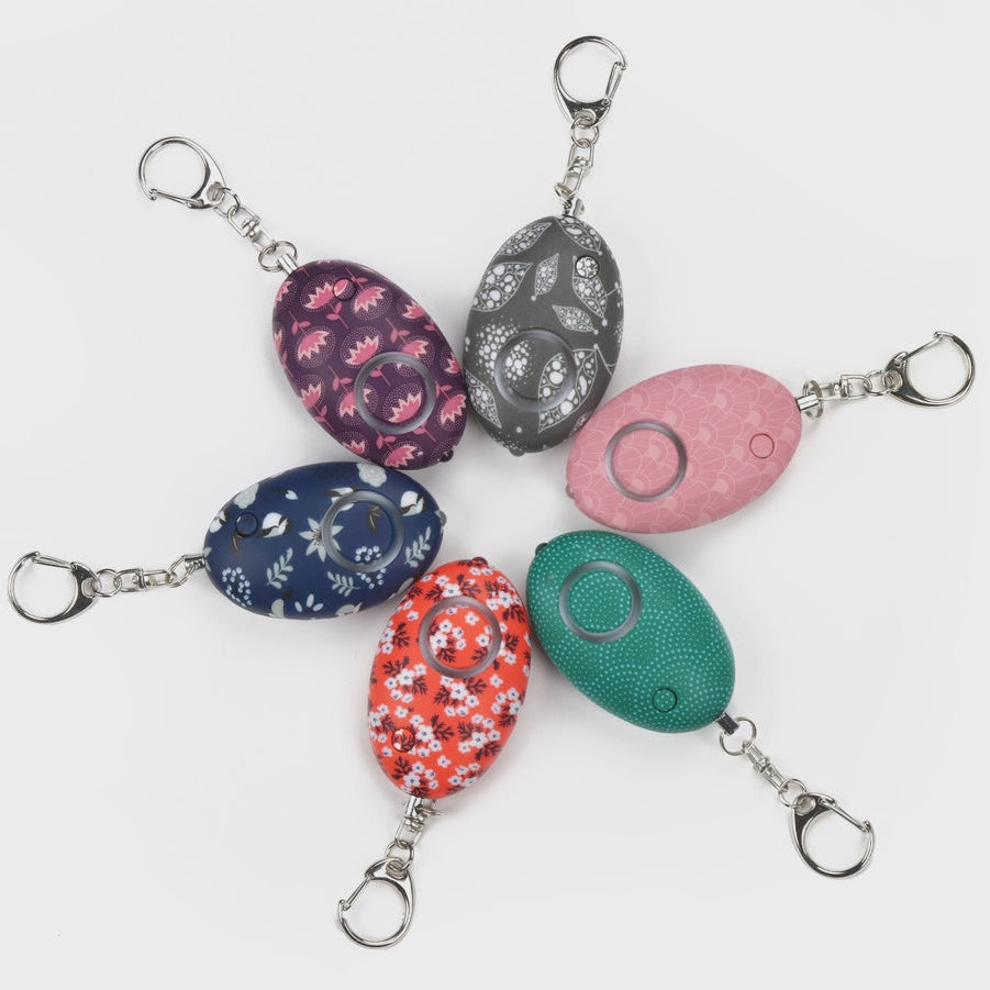 Product photograph of six patterned egg-shaped personal safety alarms arranged in a radial layout on a white background, featuring colorful designs like pink scallops and orange florals with silver carabiner keychains and small LED lights