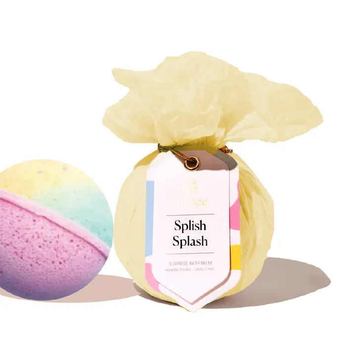 Commercial product photograph by Musee of Splish Splash surprise bath balms on a white background showcasing one textured sphere with pastel rainbow layers and a second unit wrapped in ruffled yellow tissue paper with a colorful branded tag