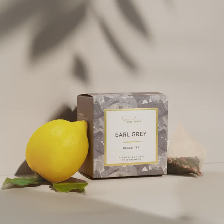 Digital lifestyle photograph by Good Provisions featuring a floral-patterned box of Earl Grey tea positioned beside a bright yellow lemon and a mesh pyramid tea bag under soft tropical leaf shadows on a neutral beige surface