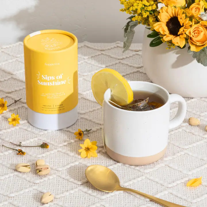 Commercial still life photograph by Happytea of a yellow Sips of Sunshine tea canister and a white ceramic mug with a lemon slice on a textured white tablecloth surrounded by vibrant sunflowers, a golden spoon, and scattered pistachios