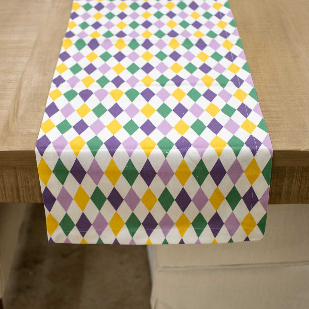 Fabric Mardi Gras table runner featuring a vibrant harlequin pattern of purple, green, and gold diamonds on a white background draped over a natural wooden table edge