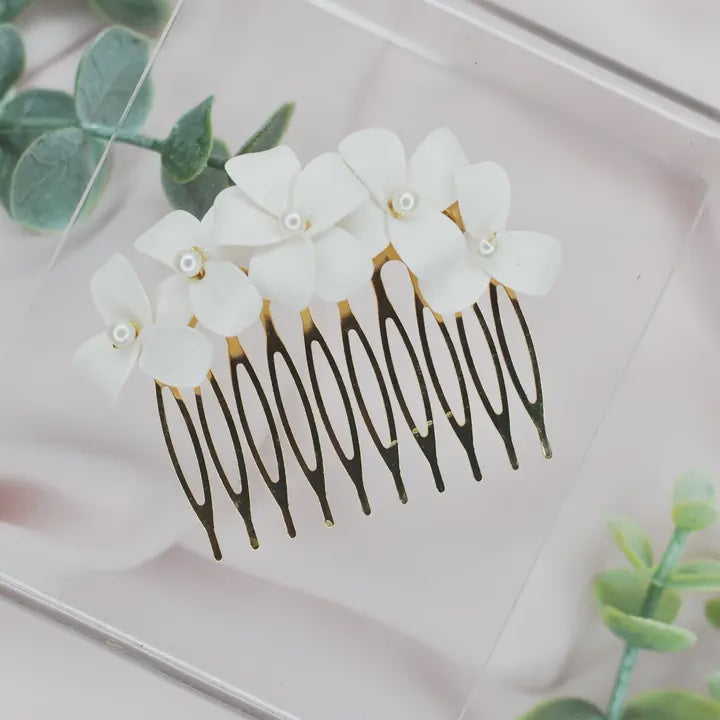 Handmade polymer clay bridal hair comb featuring delicate white four-petal flowers with central pearl accents on gold-toned metal tines resting on a clear block against a blurred green leaf background