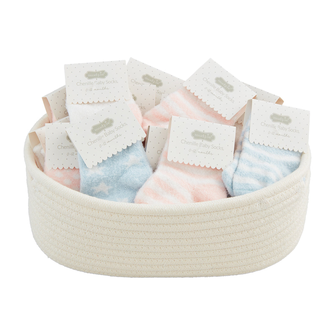 Commercial product photograph by Mud Pie showcasing an assortment of pastel pink striped and light blue star-patterned chenille baby socks tucked into a cream woven rope basket with decorative scalloped Mud Pie brand tags