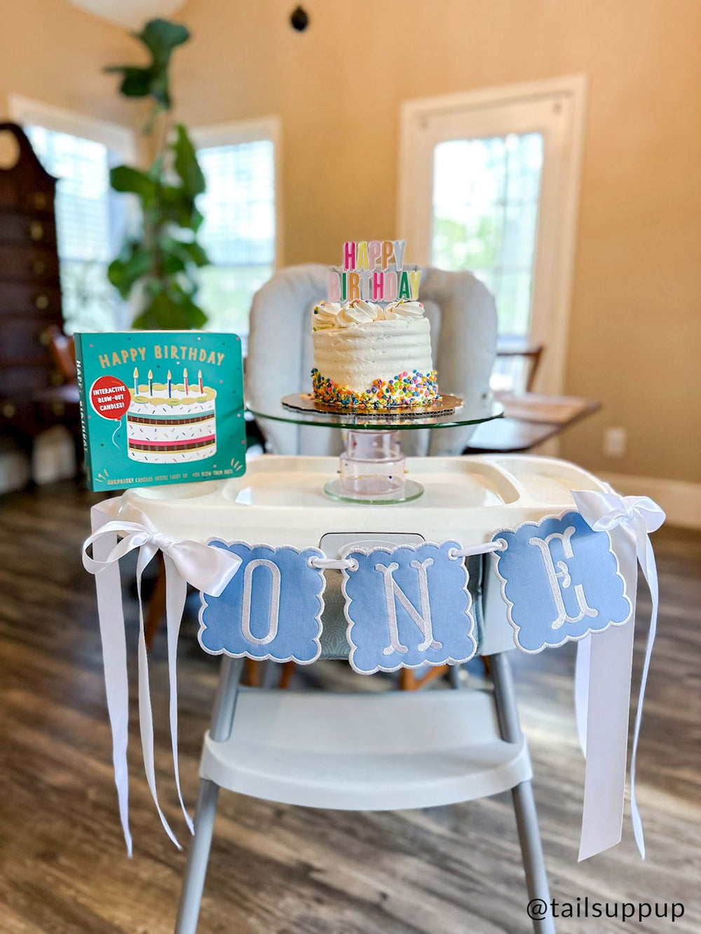 Digital photograph by tailsuppup of a first birthday party setup in a home featuring a white high chair decorated with a blue scalloped ONE banner and long white ribbons, holding a white smash cake with colorful sprinkles and a Happy Birthday book beside a fiddle leaf fig plant