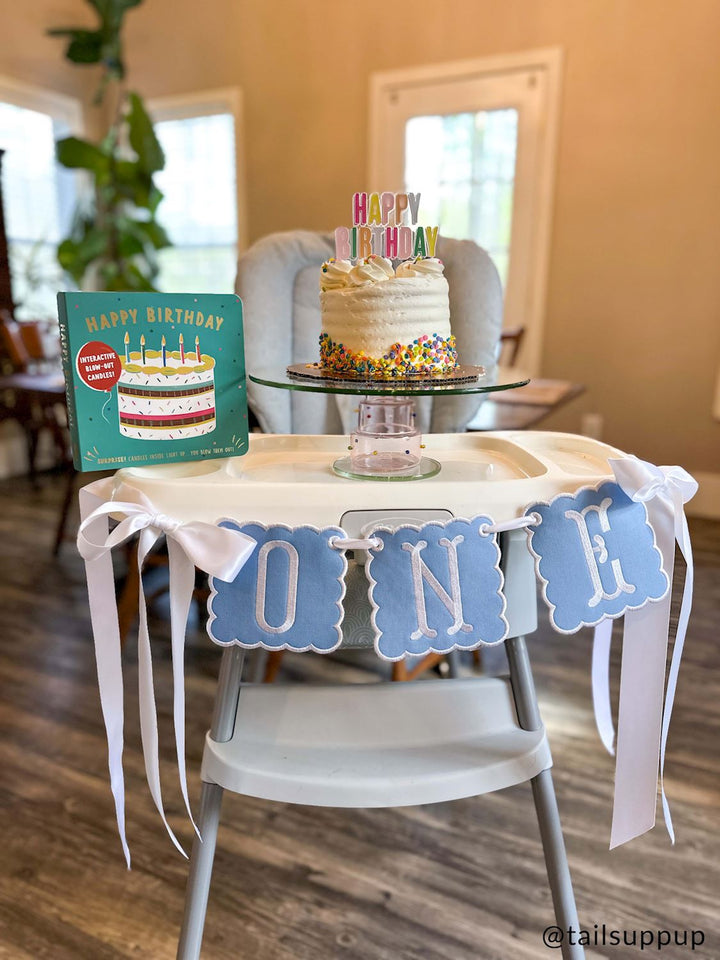 Photograph by @tailsuppup of a first birthday celebration setup on a high chair featuring a white cake with colorful sprinkles and a teal book next to a light blue scalloped ONE banner with white ribbons