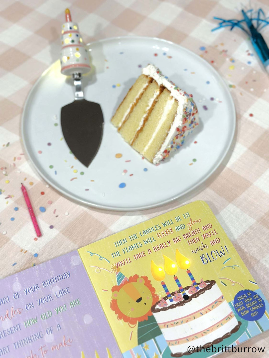 Photograph by @thebrittburrow of a four-layer vanilla sprinkle cake slice on a white plate beside an interactive birthday book and a cake server with a ceramic candle handle on a pink checkered tablecloth scattered with colorful confetti and a single pink candle