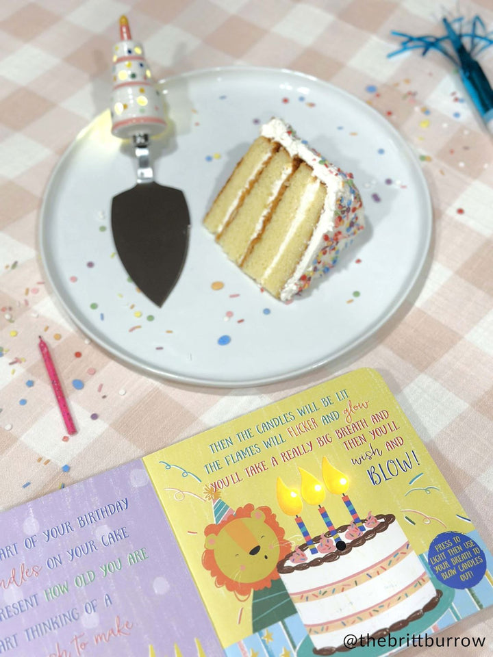 Photograph by @thebrittburrow of a four-layer vanilla sprinkle cake slice on a white plate beside an interactive birthday book and a cake server with a ceramic candle handle on a pink checkered tablecloth scattered with colorful confetti and a single pink candle