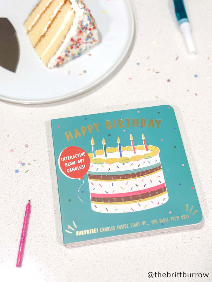 Photograph by @thebrittburrow of a teal Happy Birthday interactive board book with gold foil lettering on a white speckled surface next to a slice of sprinkle-coated vanilla cake and a pink glittery candle
