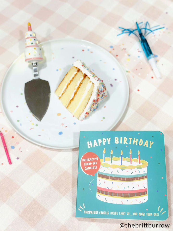 Photograph by @thebrittburrow of a slice of three-layered sprinkle cake on a white plate over a pink gingham tablecloth with a decorative tiered-cake server handle, a teal interactive birthday book, and a blue party blower