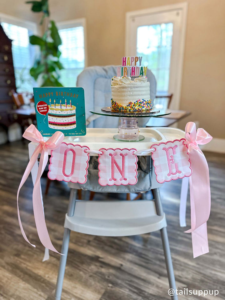 Photograph by @tailsuppup of a first birthday celebration setup on a high chair in a home featuring a white cake with rainbow sprinkles and a pink gingham ONE banner with large satin bows next to a teal interactive book