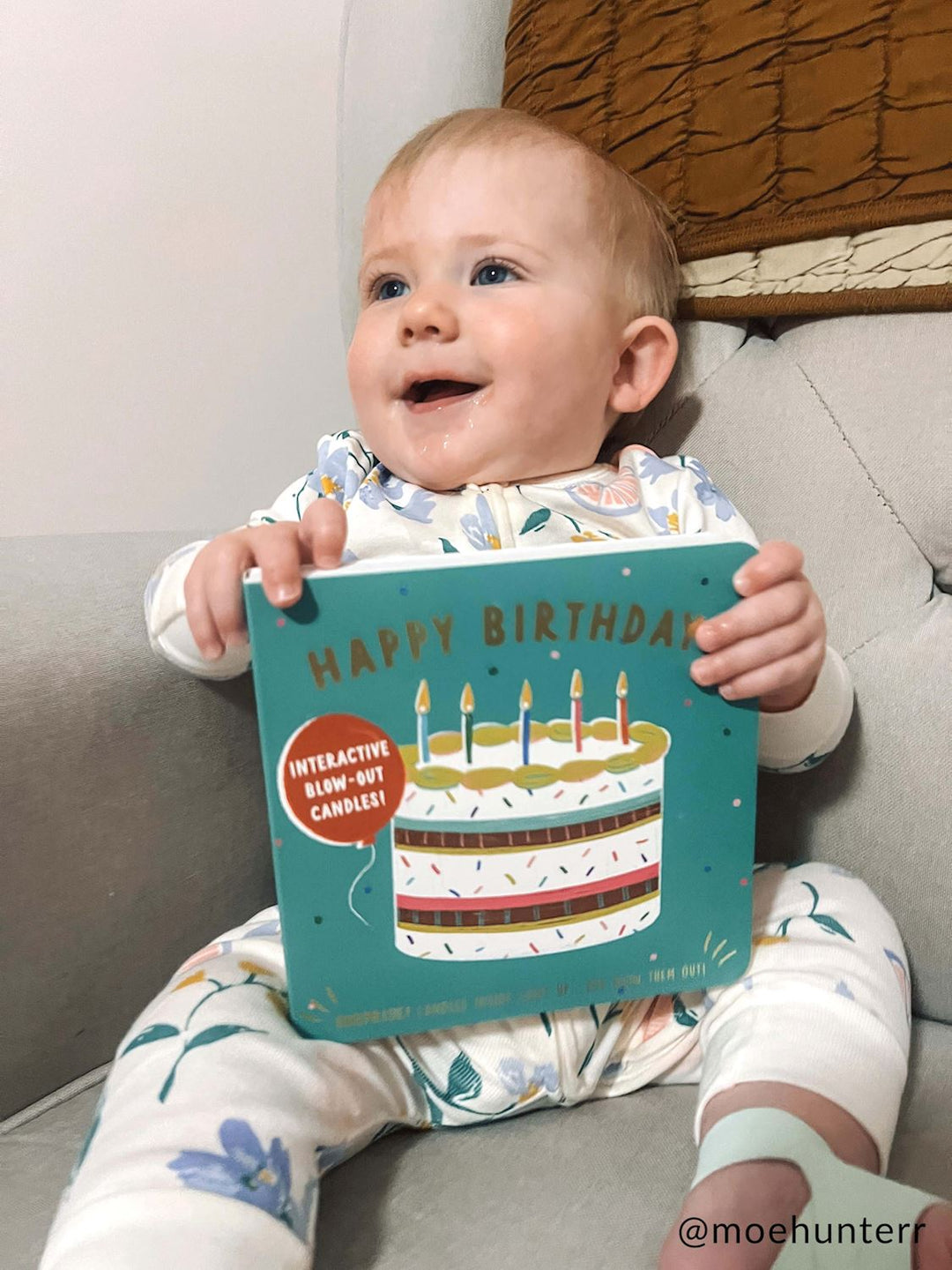 Digital photograph by @moehunterr of a smiling blue-eyed baby sitting on a grey sofa holding a teal Happy Birthday board book with an interactive cake illustration while wearing white floral-print pajamas