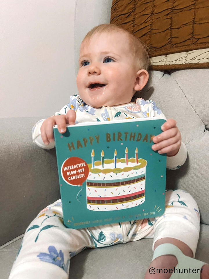 Digital photograph by @moehunterr of a smiling blue-eyed baby sitting on a grey sofa holding a teal Happy Birthday board book with an interactive cake illustration while wearing white floral-print pajamas