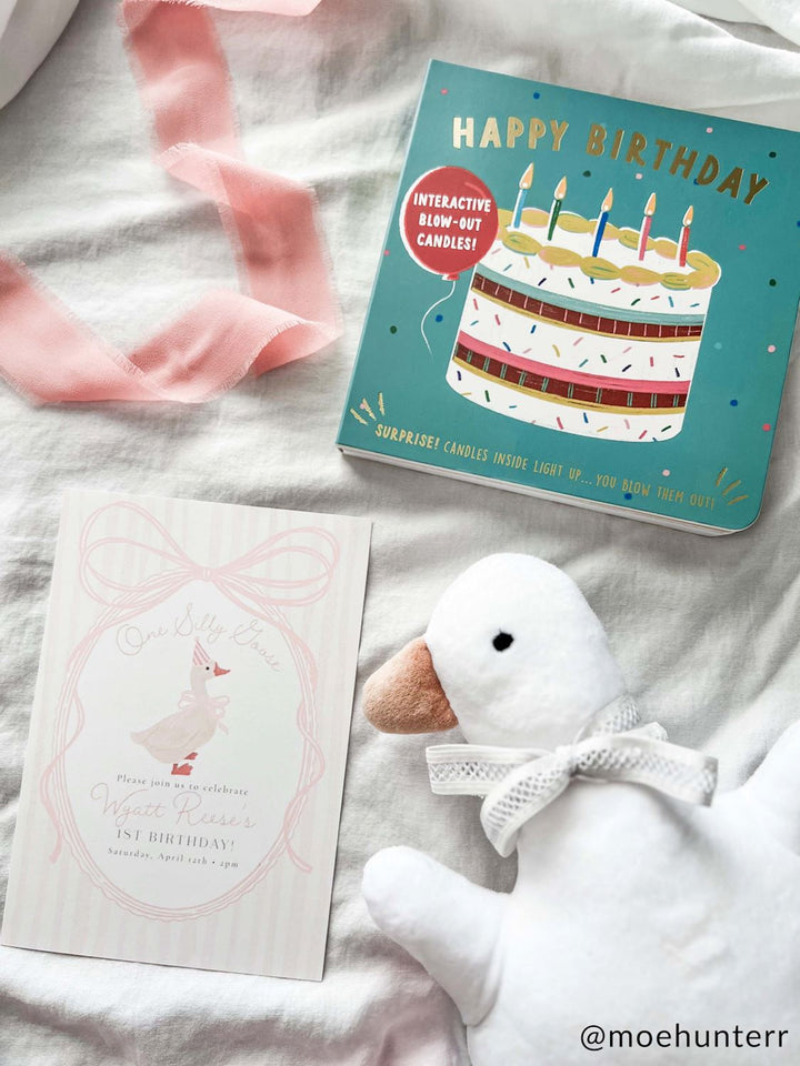 Photograph by @moehunterr of a first birthday flat lay on white fabric featuring a teal interactive Happy Birthday book, a One Silly Goose invitation card, and a white plush goose wearing a lace bow