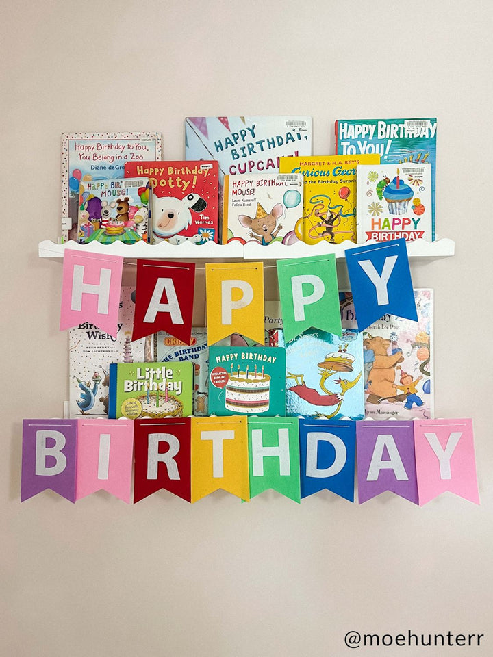 Photograph by @moehunterr of birthday-themed children’s books including Dr. Seuss and Curious George on white wall shelves featuring a multi-colored felt pennant banner and bright illustrations of cakes and party hats