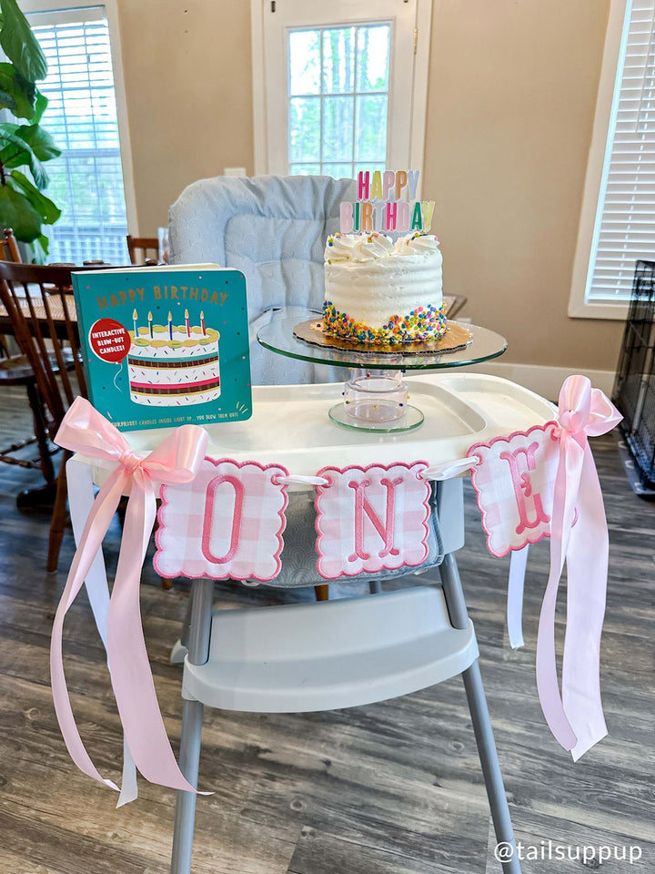 Digital photograph by @tailsuppup of a first birthday celebration setup in a home dining area featuring a white sprinkle-covered cake on a high chair decorated with a pink gingham ONE banner and large satin bows next to a teal interactive birthday book