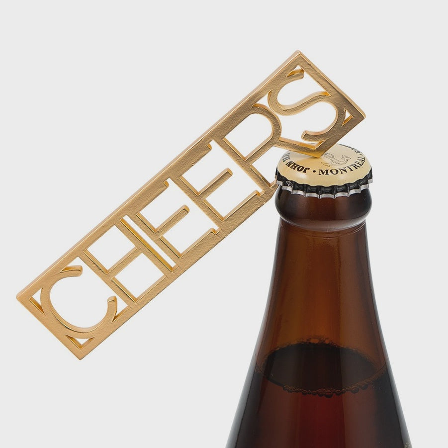 Cast brass bottle opener shaped like the word Cheers is poised on a brown glass beer bottle in a minimalist studio setting, showcasing a reflective gold-toned finish, elegant serif typography, and a white bottle cap featuring Montreal branding