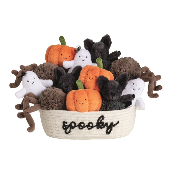 Photograph of Jellycat Halloween plush toys featuring smiling orange pumpkins, fuzzy grey bats, and white ghosts nestled inside a white woven rope basket embroidered with the word spooky in black cursive lettering