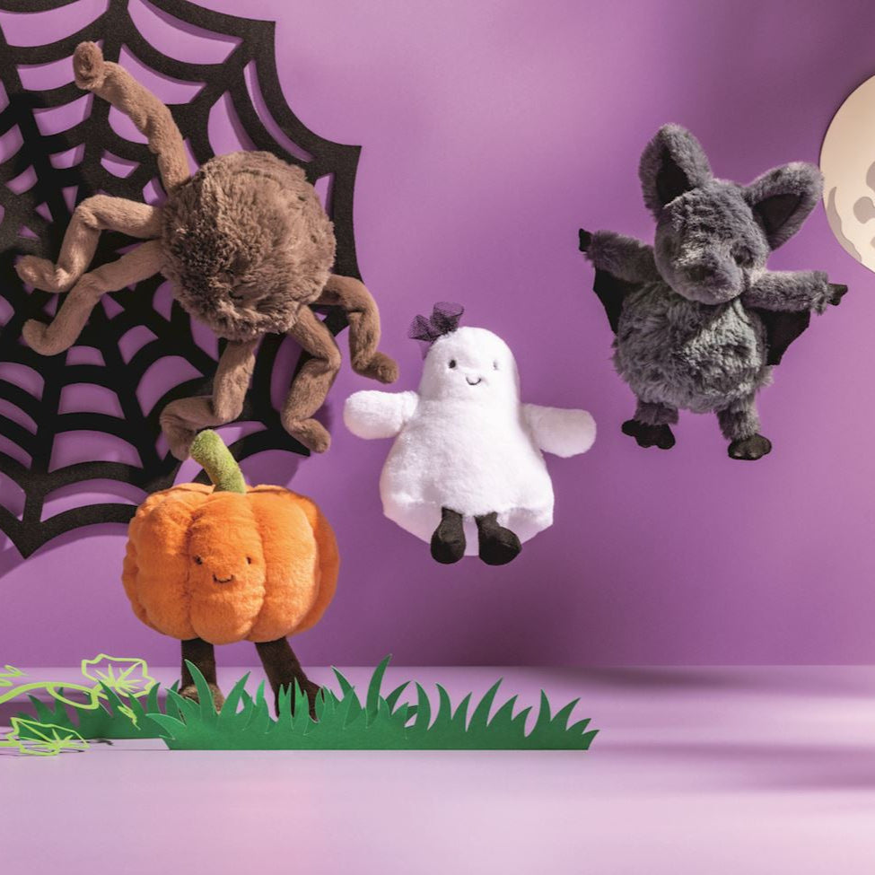 Plush stuffed toys by Jellycat featuring Halloween characters including a smiling orange pumpkin, a white ghost with a tiny purple bow, and a fluffy grey bat against a purple backdrop with a black spider web and green paper grass
