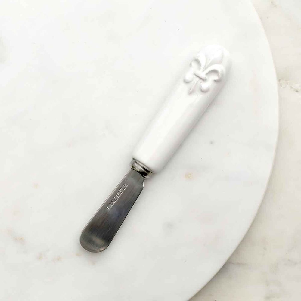 Stainless steel and ceramic cheese spreader resting on a white marble plate featuring a white handle with a raised fleur-de-lis motif and a rounded silver blade engraved with the words stainless steel