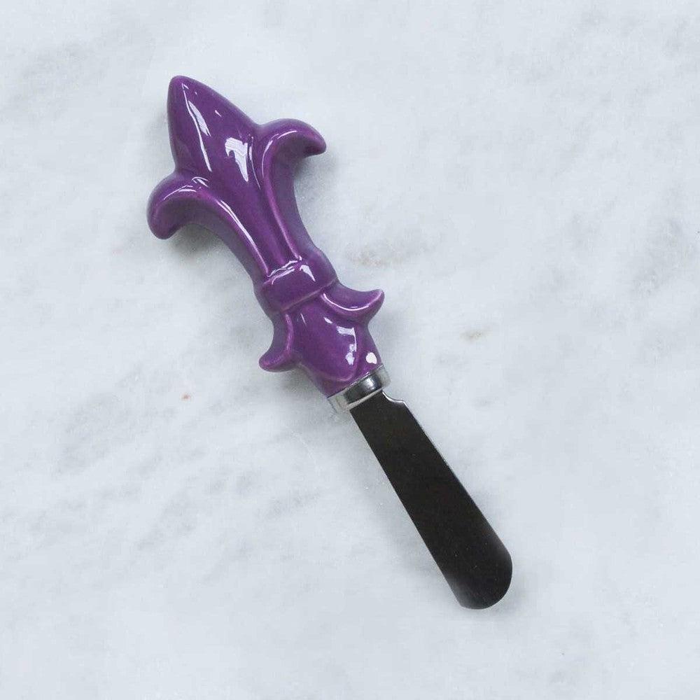 Ceramic and stainless steel cheese spreader with a vibrant purple fleur-de-lis handle on a white marble surface featuring a glossy finish and a rounded metal blade