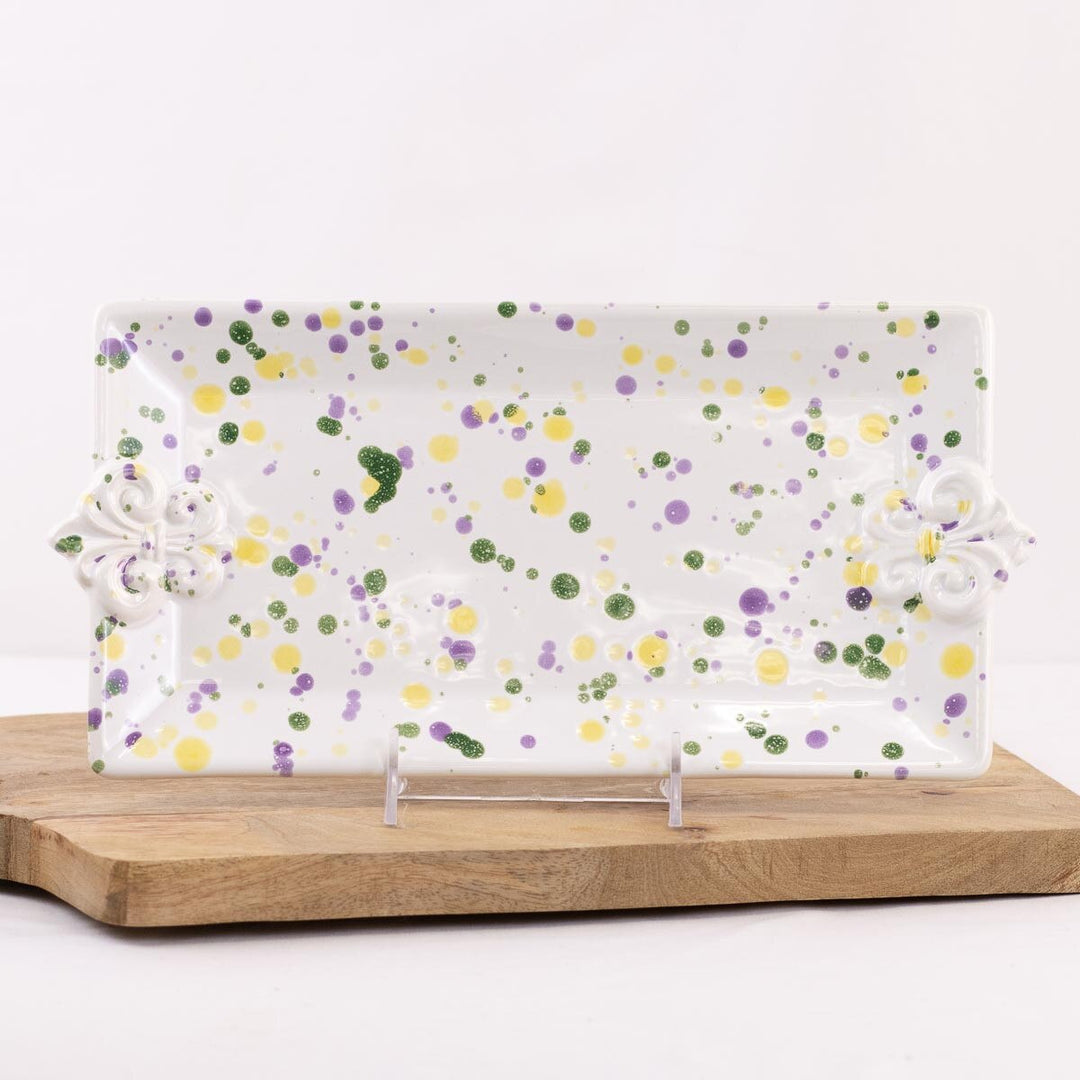 Ceramic rectangular serving platter featuring a festive purple, green, and yellow splatter pattern and raised fleur-de-lis handles resting on a natural wooden block against a white backdrop
