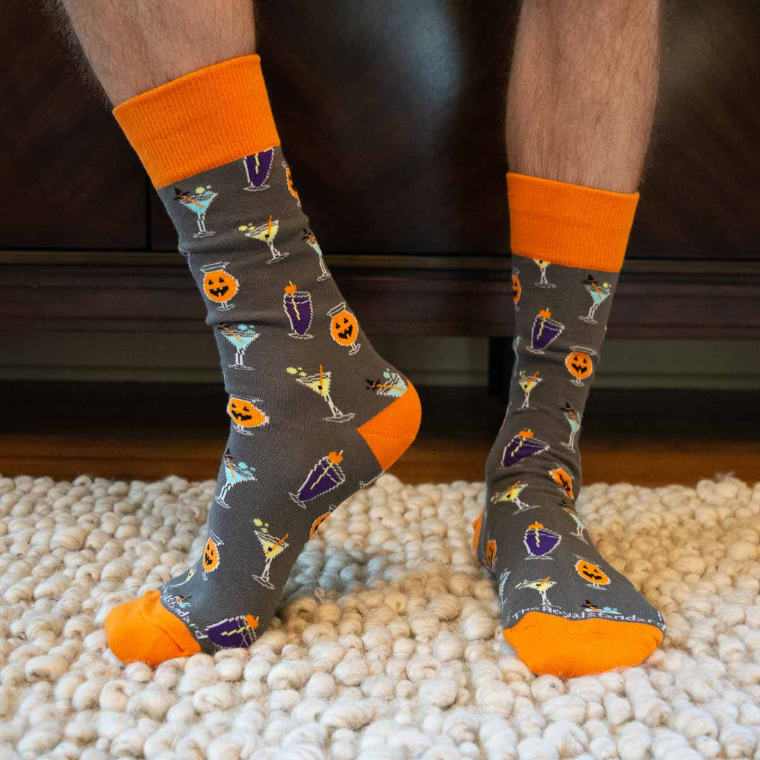 Digital photograph of The Royal Standard socks worn by a person on a white shag rug featuring a grey background with bright orange accents and a pattern of jack-o'-lantern cocktail glasses and purple drinks with witch hats