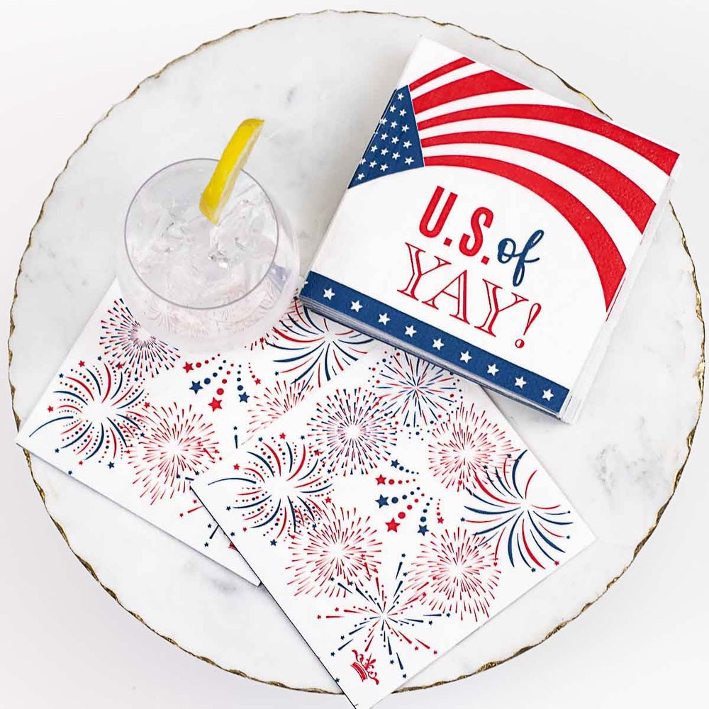 Lifestyle photograph of patriotic paper napkins on a gold-rimmed marble platter featuring the phrase U.S. of YAY and red and blue firework designs next to a glass of ice water with a yellow lemon wedge