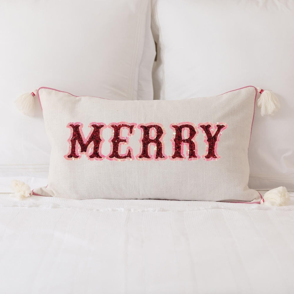 Photograph of a decorative lumbar pillow featuring the word MERRY on a white bed with red sequined lettering, pink piping, and white tassels at each corner