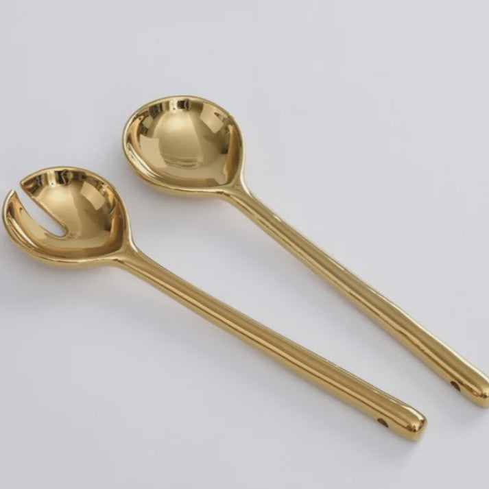 Polished gold-toned metal salad servers by an unidentified designer resting on a neutral white background featuring a brilliant mirror-like reflection, elongated cylindrical handles, and small hanging holes at the base