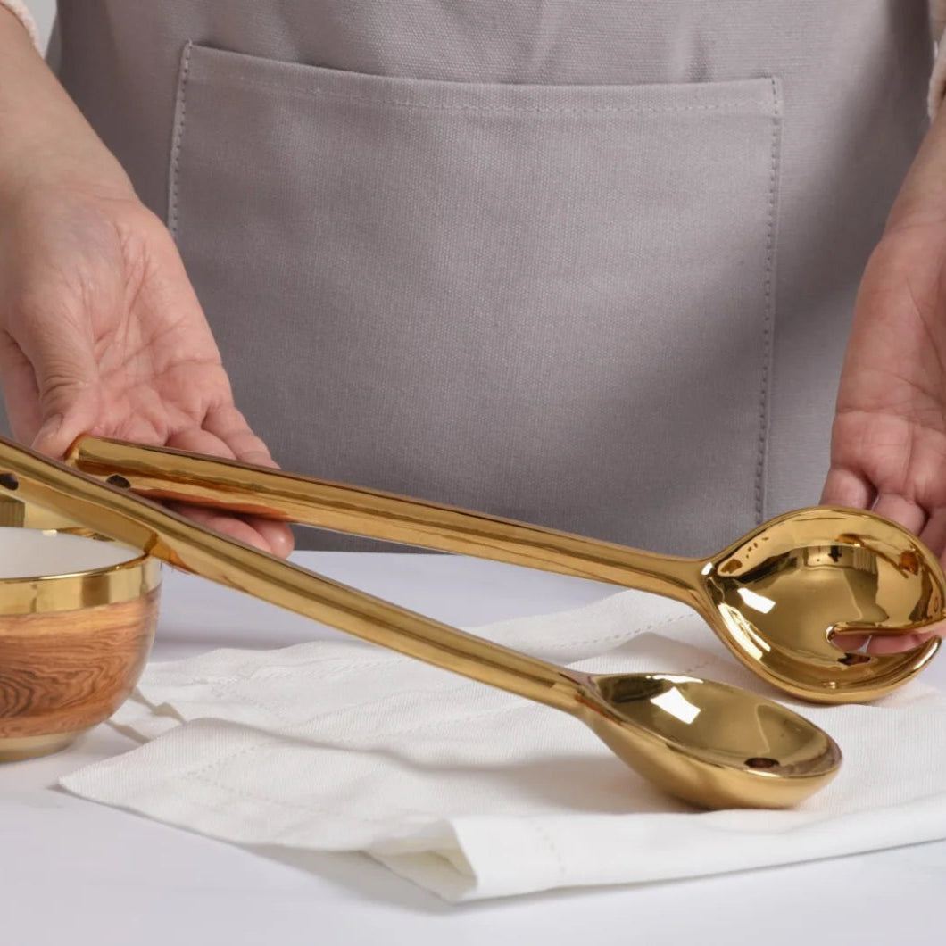 Polished gold-finished stainless steel salad servers held by a person wearing a grey apron over a white cloth napkin beside a matching gold-rimmed wooden bowl