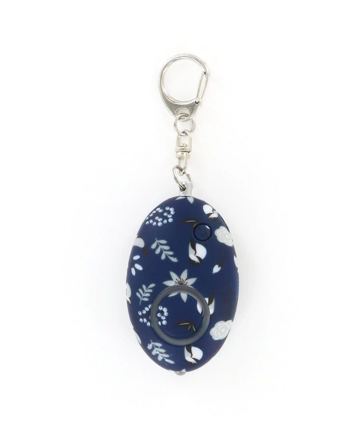 Personal safety alarm by Birdie featuring a navy blue oval-shaped device centered on a white background with a white floral and bird pattern, a silver metal lobster clasp, and a circular activation button