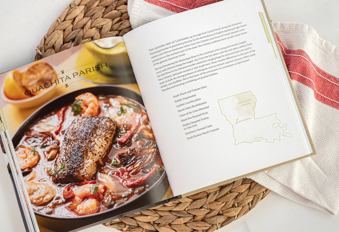 Photograph of an open cookbook highlighting the culinary heritage of Ouachita Parish, Louisiana, featuring a rich catfish courtbouillon with shrimp next to a gold map outline and a red-striped kitchen towel on a woven mat