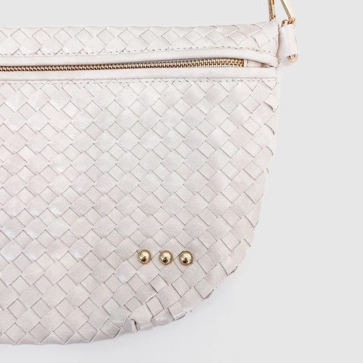Photograph of a cream woven half-moon handbag on a white background featuring a gold-tone zipper, three decorative metallic studs, and an intricate braided texture