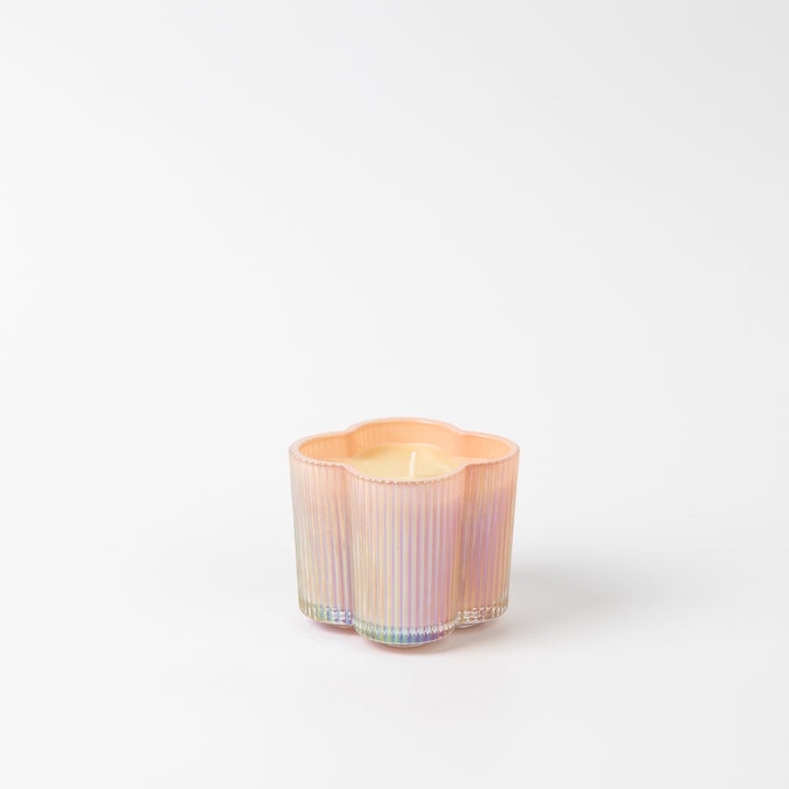 Studio photograph of a peach iridescent glass candle featuring a vertical ribbed texture and a scalloped clover shape filled with cream-colored wax on a white background